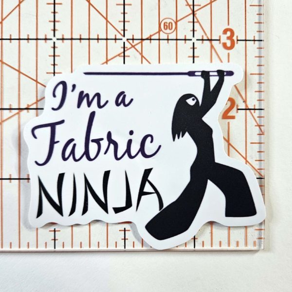 Sticker with I'm a fabric Ninja written next to a ninja outline of a woman with long hair , white mask, and wielding a large sewing needle.