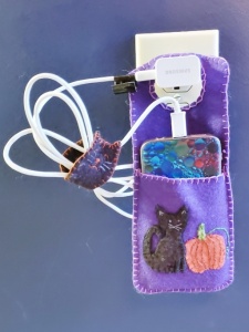 Purple felt cell phone charging pouch with black cat and orange pumpkin plugged into and hanging from wall.