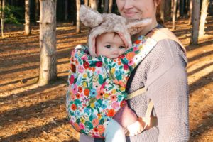 Brightly flowered baby carrier with brown haired child wearing furry hood. Parent is standing in the woods