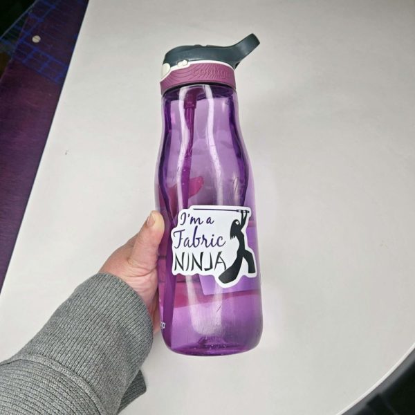 Sticker with I'm a fabric Ninja written next to a ninja outline of a woman with long hair , white mask, and wielding a large sewing needle. on a purple water bottle