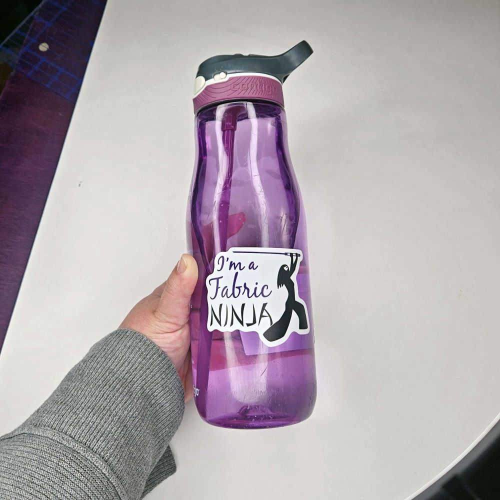 Sticker with I'm a fabric Ninja written next to a ninja outline of a woman with long hair , white mask, and wielding a large sewing needle. on a purple water bottle