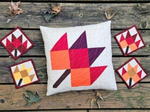 Quilted Maple leaf pillow cover and coasters made from reds, yellows and oranges on a rustic wood background