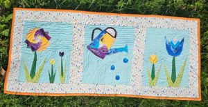 Small quilt laying on the grass. Quilt has a purple and yellow flower, a watering can, and a 2 tulips, blue and yellow.