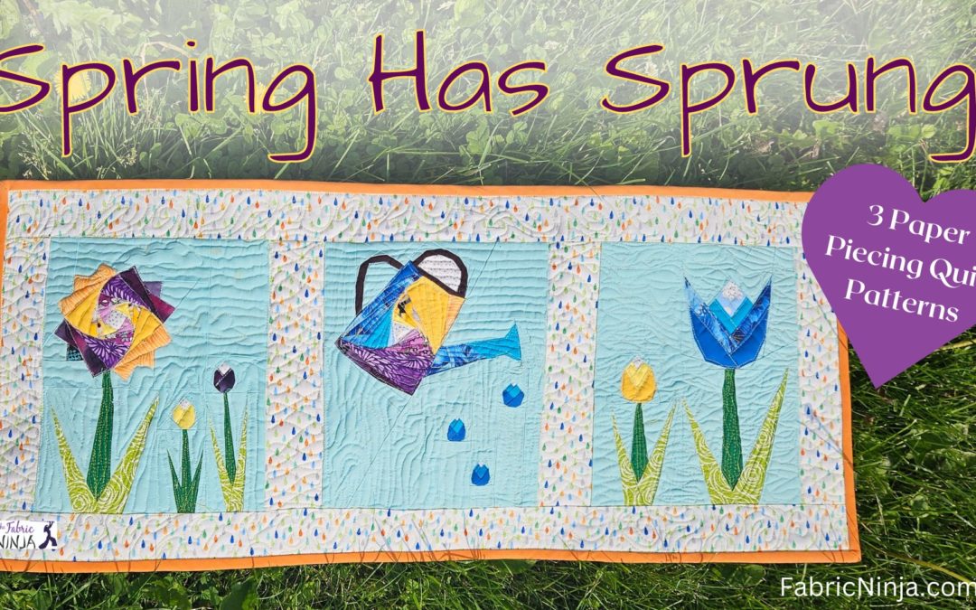 Small quilt laying on the grass. Quilt has a purple and yellow flower, a watering can, and a 2 tulips, blue and yellow.