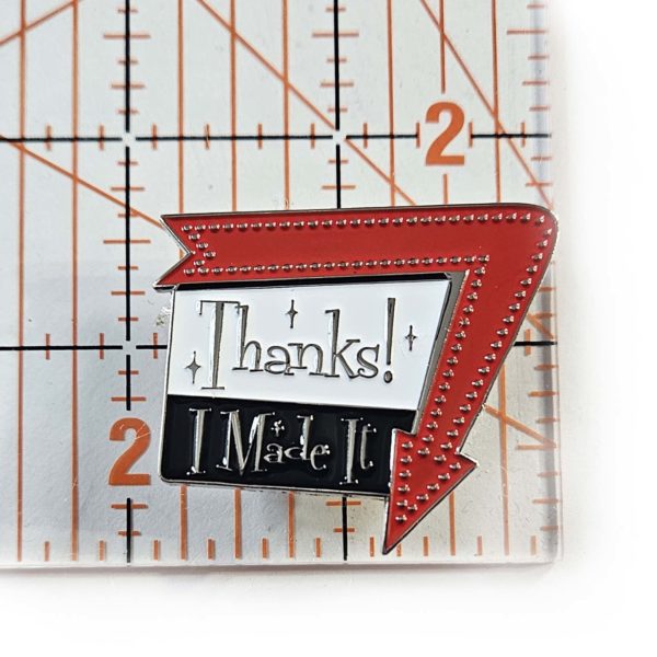 Enamel pin says, "Thanks! I Made It" in a 1960 sign style with arrow around one edge pointing down. Arrow is red and the sign is black and white with silver metal