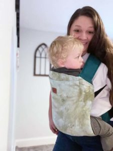 white woman with dark hair wearing blond haired baby in a happy baby toddler carrier with a green tie-dye slipcover