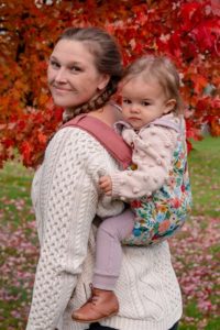 flower slipcover on pink onbuhimo happy baby carrier. Baby in carrier is wearing pastel and cream clothing has pale skin and light brown hair