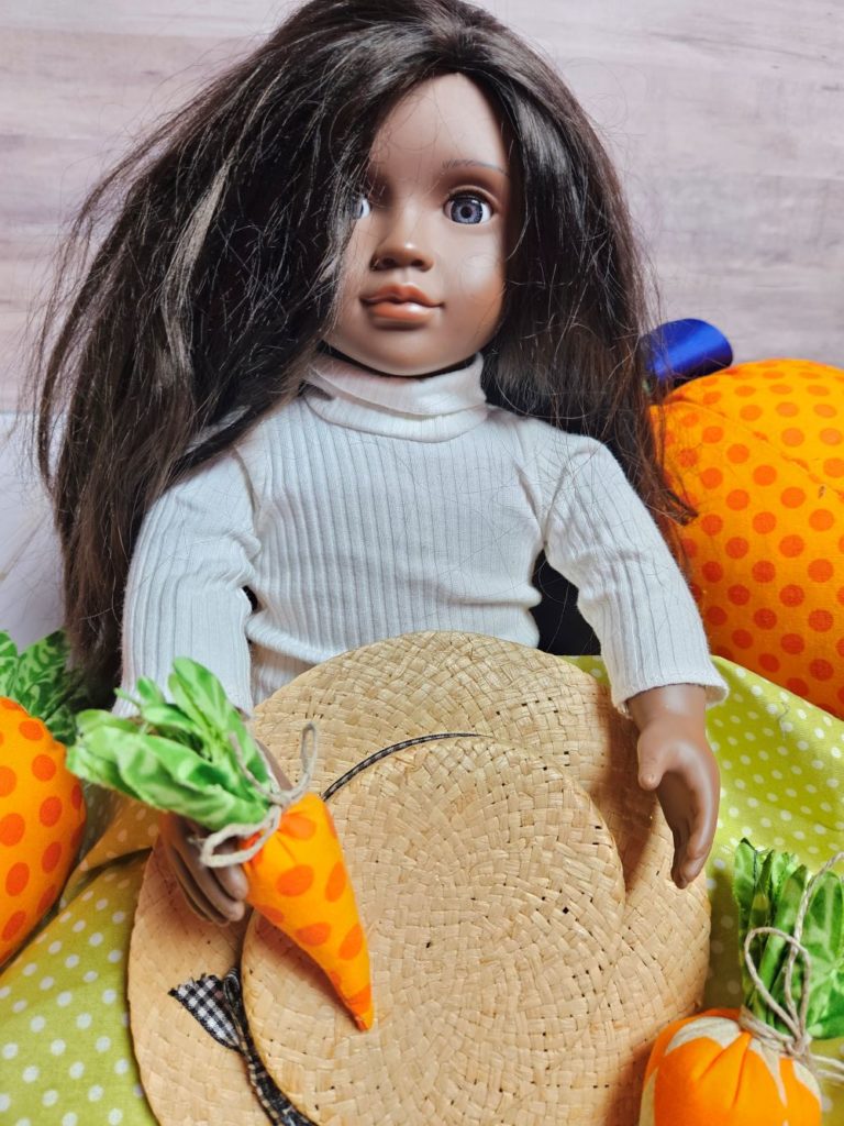doll with dark skin and hair wearing a white turtle neck sweater has a hat in her lap and is holding a fabric carrot