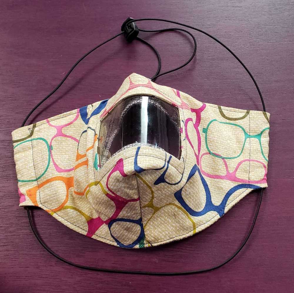 A mask made of tan fabric with colorful glasses printed on it. The mask has a clear window in the front and elastic loops around the head