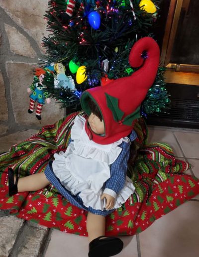 "Just add ears" elf hood being worn by doll - Christmas tree and fireplace in background.