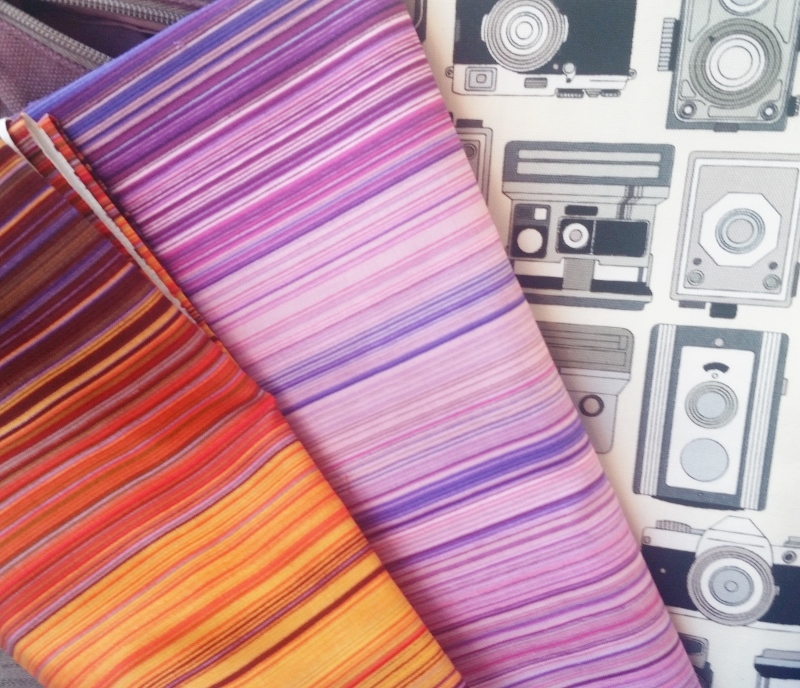Three pieces of fabric. Shades of orange stripe, shade of pink and purple stripe, and black and white cameras