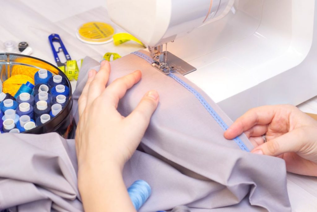 White hands sewing grey fabric on a sewing machine.