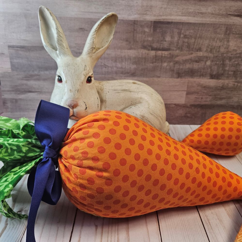 Large orange polka dotted stiffed carrot with blue bow in front of a white rabbit sculpture