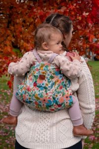 flower slipcover on pink onbuhimo happy baby carrier. Baby in carrier is wearing pastel and cream clothing has pale skin and light brown hair