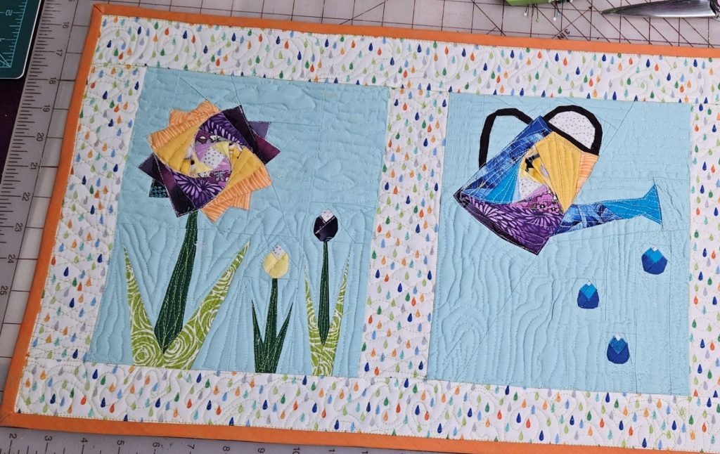A flower and watering can quilt. Body of watering can and the flower head are made of a fabric swirl pattern with yellow, blue and purple 