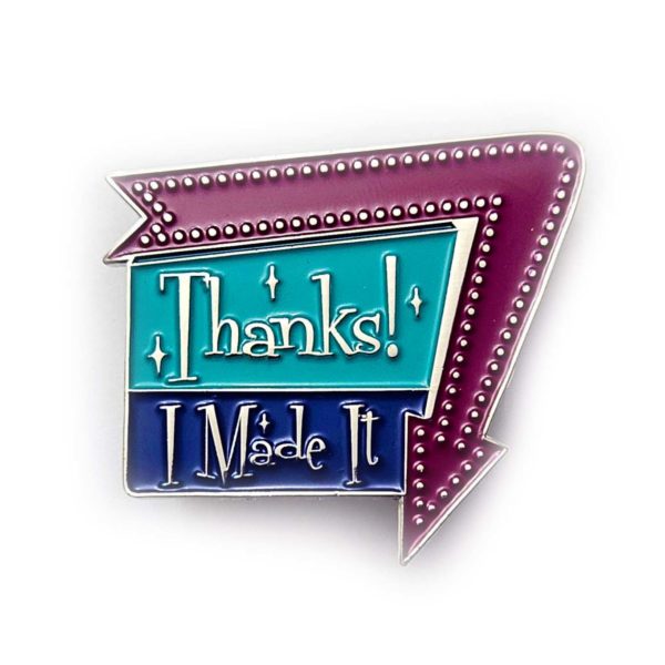 Enamel pin says, "Thanks! I Made It" in a 1960 sign style with arrow around one edge pointing down. Arrow is a warm purple, and the sign is aqua and dark blue with silver metal