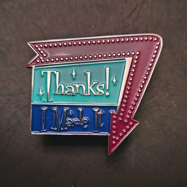 Enamel pin says, "Thanks! I Made It" in a 1960 sign style with arrow around one edge pointing down. Arrow is a warm purple, and the sign is aqua and dark blue with silver metal