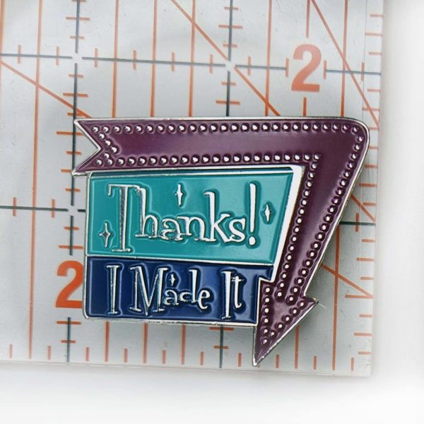 Enamel pin says, "Thanks! I Made It" in a 1960 sign style with arrow around one edge pointing down. Arrow is a warm purple, and the sign is aqua and dark blue.