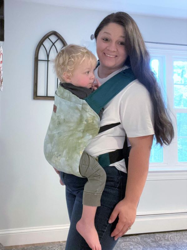 White woman with dark hair with toddler on her front in a Happy Baby Toddler carrier. It is green the straps are teal. 