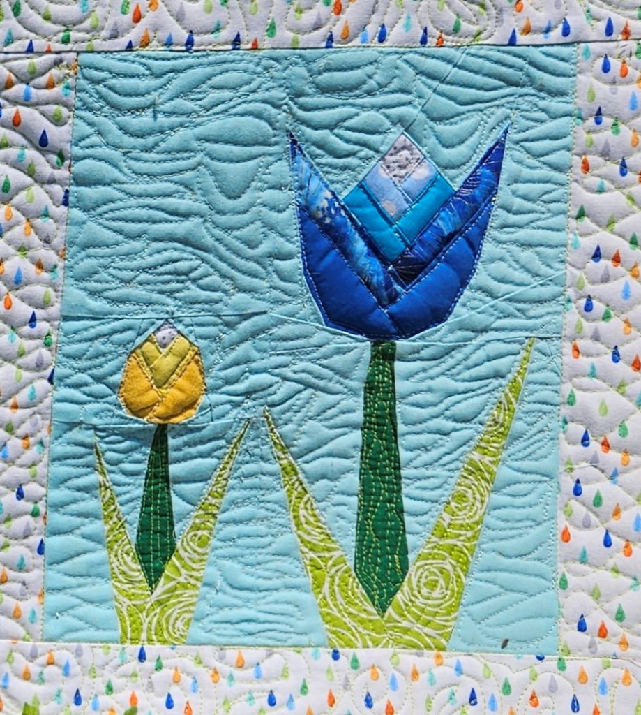two tulips in a quilt block. a large blue one with several shade of blue and a white tip. Then a small yellow bud, both with green leaves on a light blue quilted background