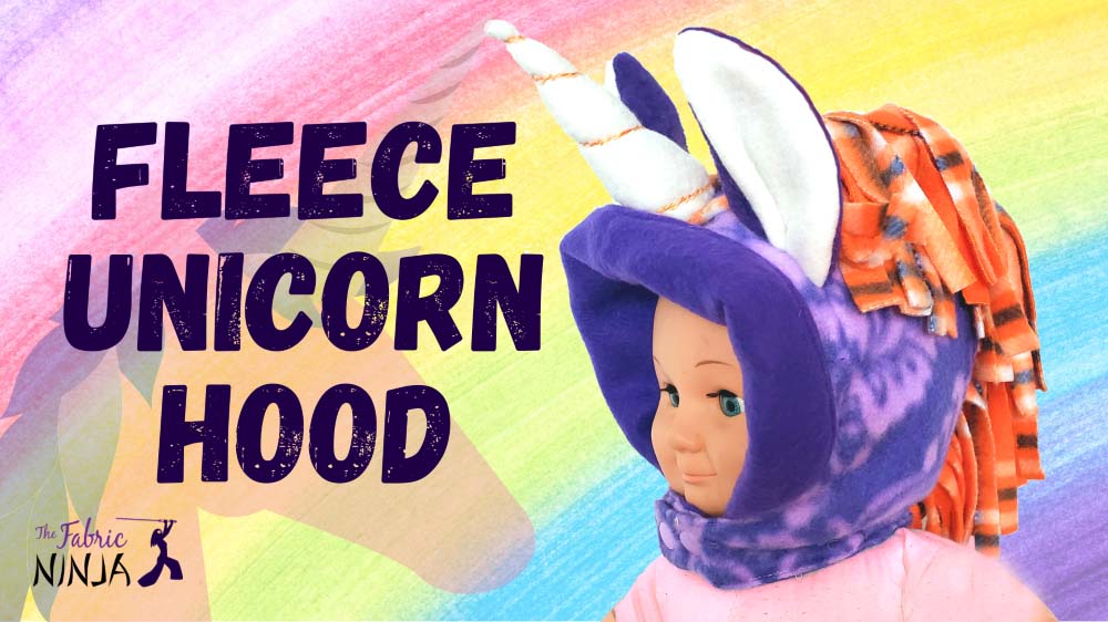 Fleece Unicorn Hood rainbow background. Doll wearing a purple unicorn hood with orange mane and white horn