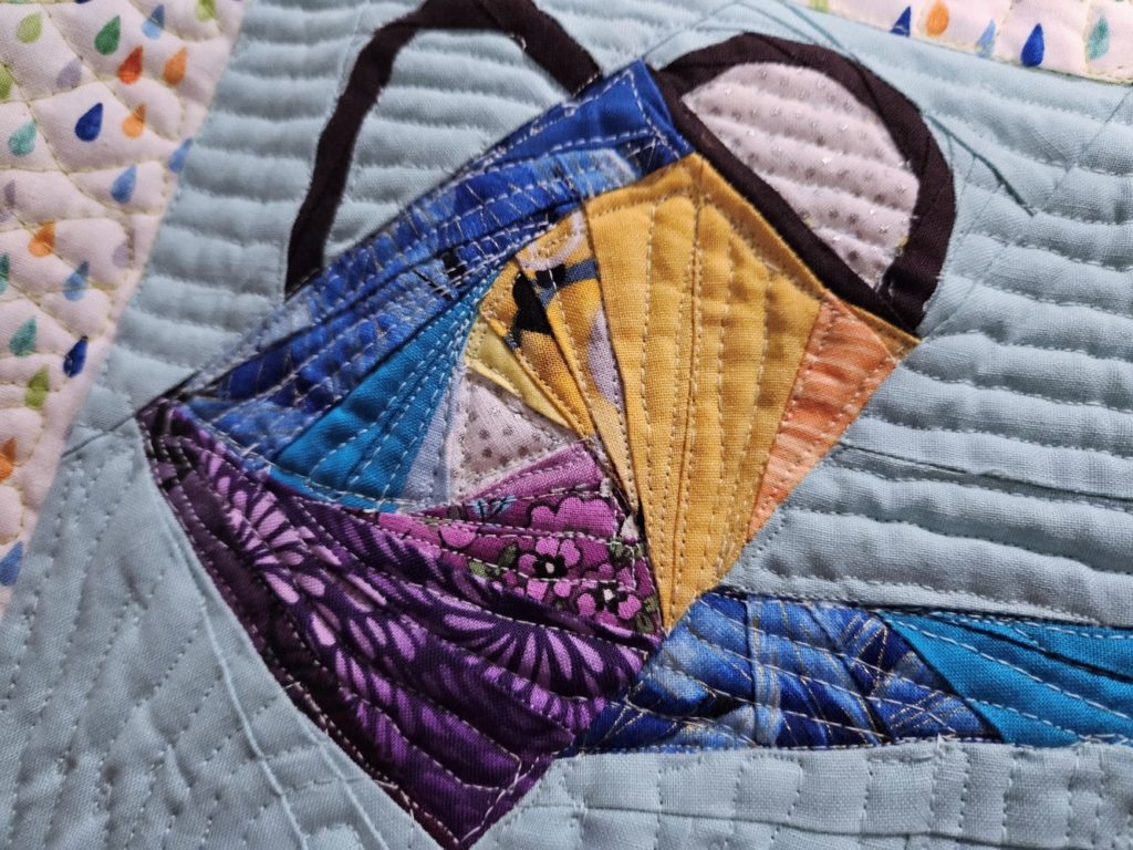 close up of watering can quilt block. Body of can is in a swirl pattern with yellow, blue and purple swirls around the center 