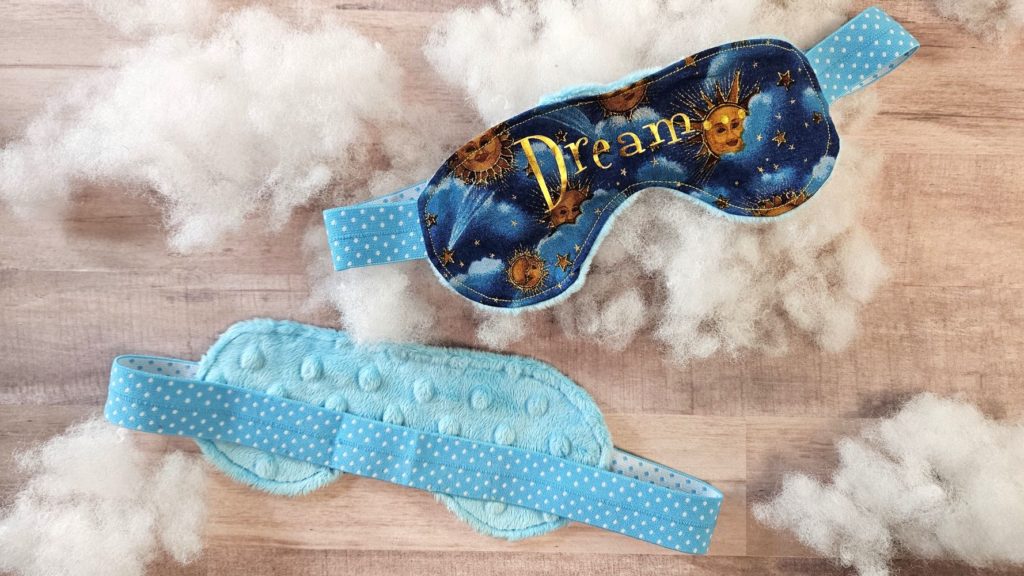 2 sleeping masks on cloud looking puffs. One eye mask says dream in gold on a dark blue mask with suns. The other is light blue and fuzzy. 